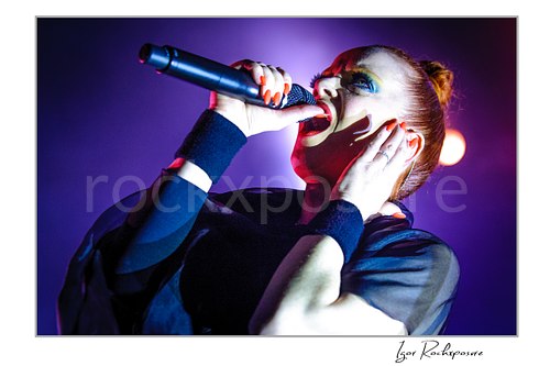 Horizontal color image of Shirley Manson singing into a microphone during a Garbage concert in an intense close-up under vivid purple and pink stage light