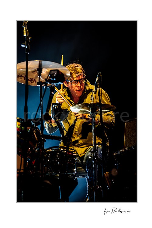 Vertical color image of Patrick Carney of The Black Keys playing drums under warm stage lighting and wearing dark glasses