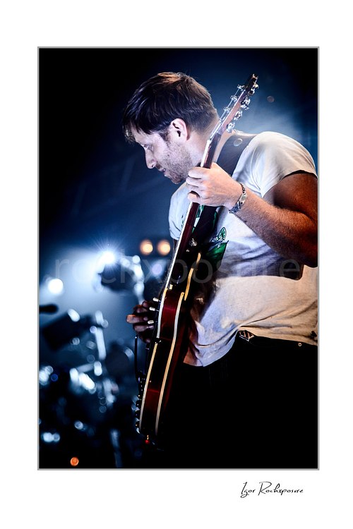 Vertical color image of Dan Auerbach of The Black Keys playing guitar in side profile under bright blue stage lights