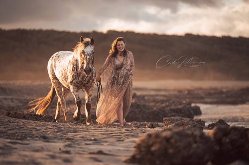 Frau in Glitzerkleid führt ihr Appaloosa-Pferd am Strand bei Sonnenuntergang