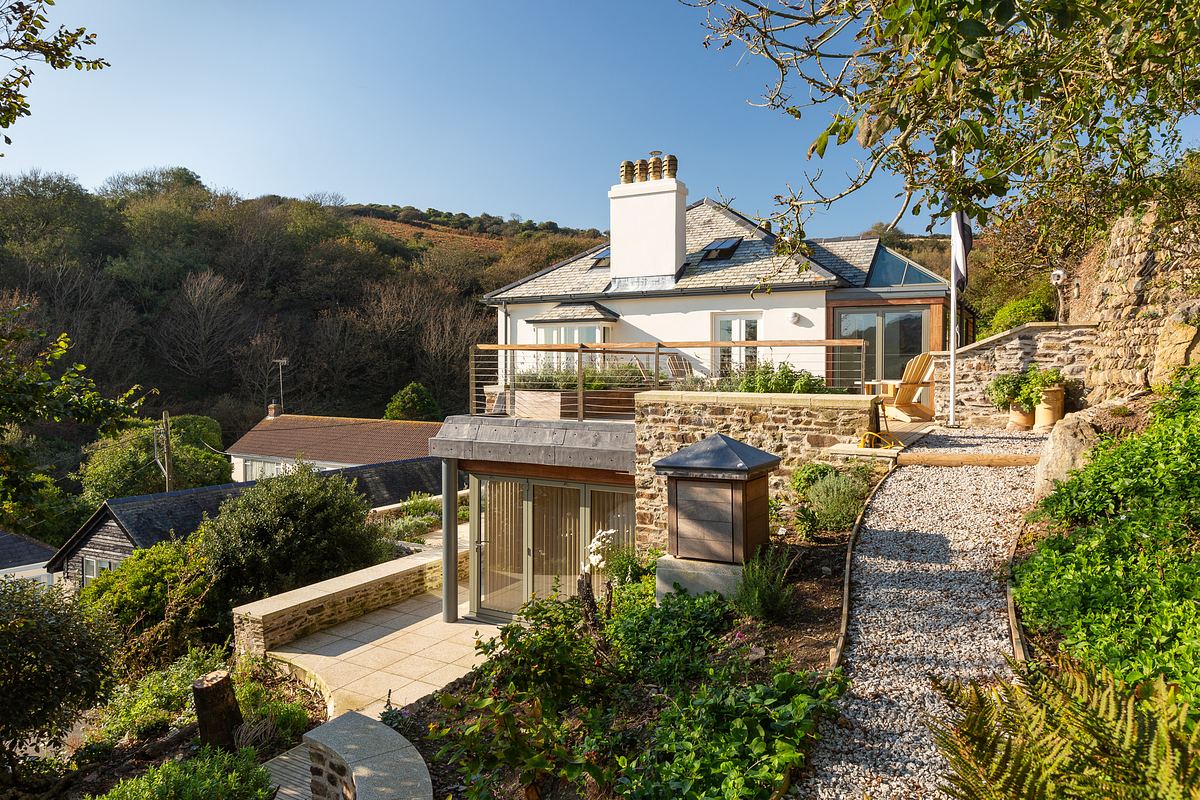 A modern house in a village in Cornwall with a stone patio and glass extension sits on a hillside, surrounded by greenery and a gravel path exterior photographed in optimal daylight.