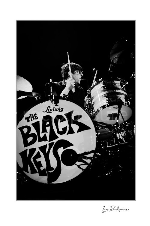 Vertical black and white image of Patrick Carney of The Black Keys playing drums beside a bass drum marked The Black Keys