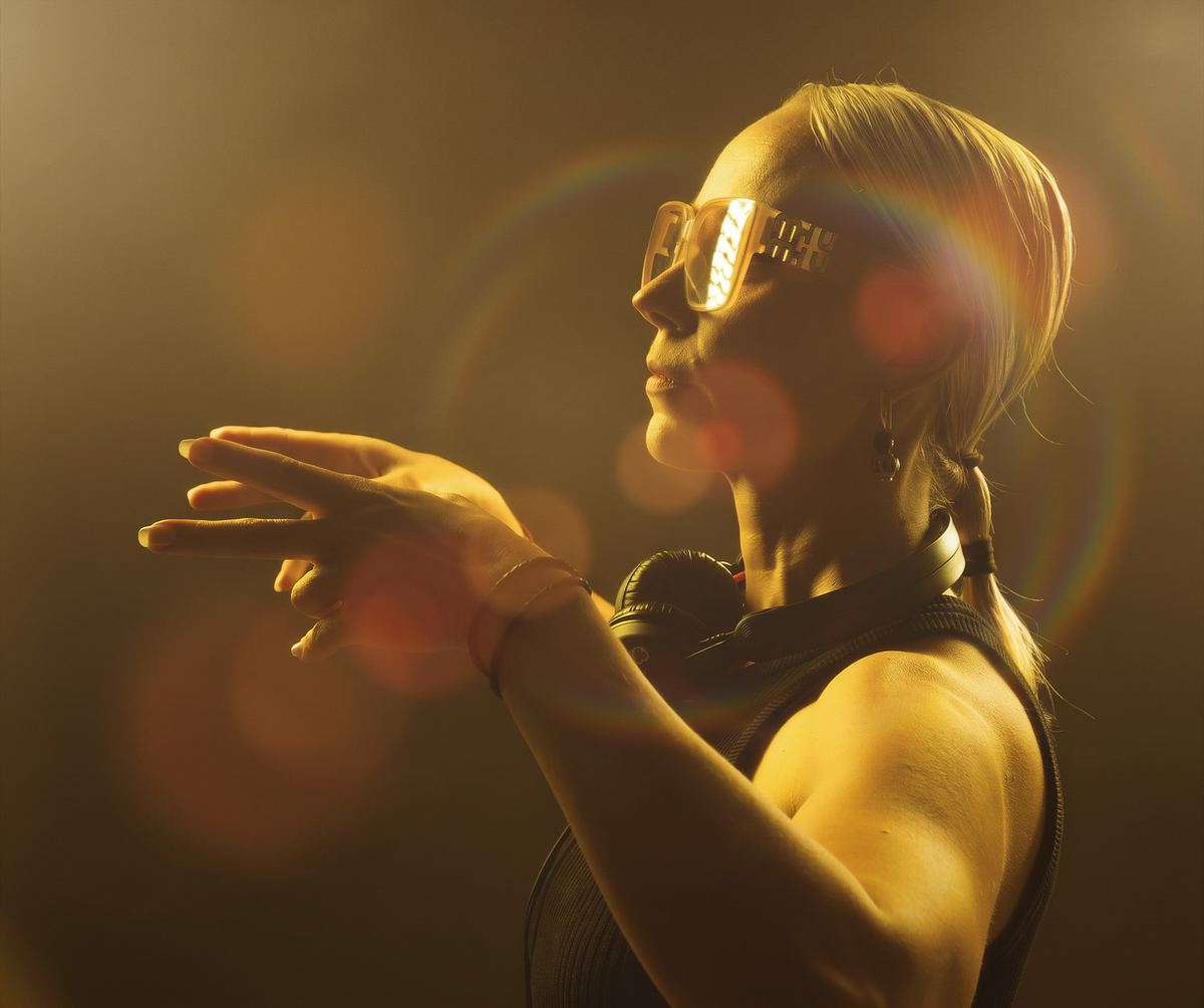 A person wearing reflective sunglasses and headphones gestures with their hands in warm, yellow lighting with lens flares.