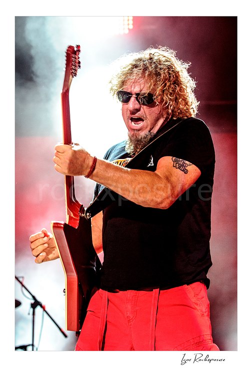 Vertical color image of Sammy Hagar playing guitar onstage in sunglasses under bright backlight and red stage haze