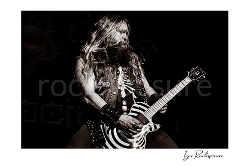 Black and white shot of Zakk Wylde wearing a denim vest, playing his signature bullseye guitar on stage against a dark background.