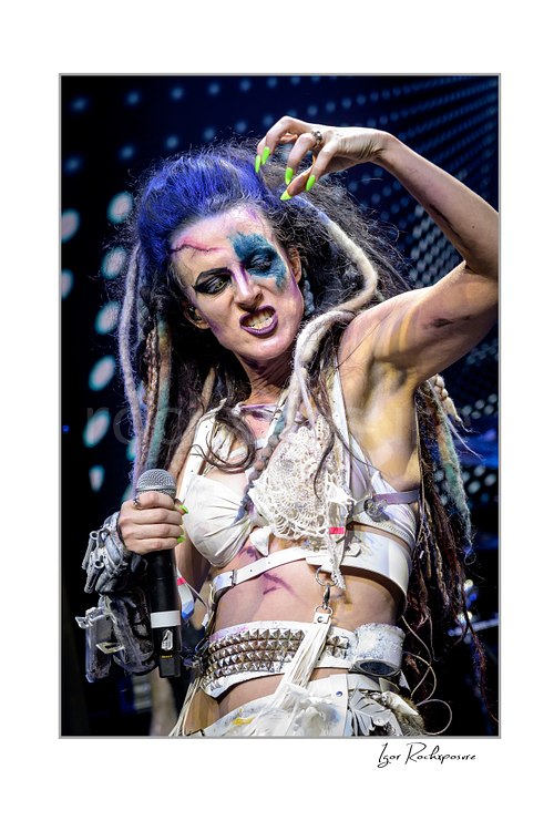 Vertical color image of Calico Cooper of Beasto Blanco holding a microphone in a fierce live performance close-up, with dramatic stage makeup, dreadlocks, and neon green nails