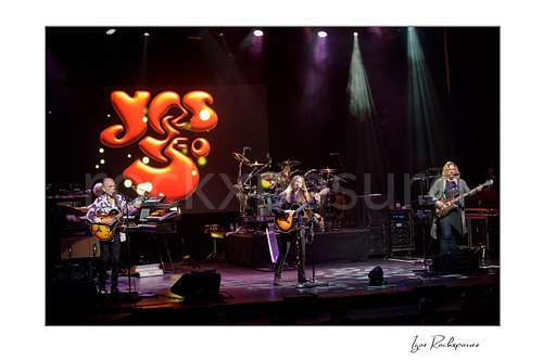 Horizontal color image of Yes performing onstage with the glowing Yes logo on the backdrop and dramatic overhead spotlights