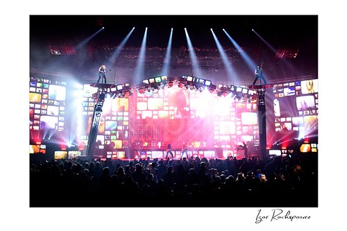 Horizontal color image of Trans Siberian Orchestra performing on stage with guitarists on raised platforms, bright video screens, and a packed arena crowd