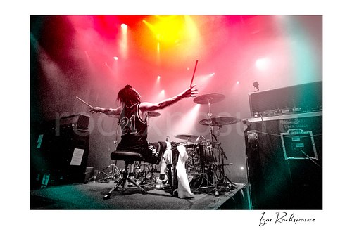 Horizontal color image of Zoltan Chaney at the drum kit with arms outstretched under red, yellow, and white stage lights