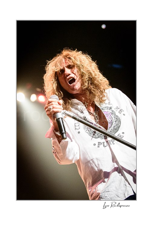 Vertical color image of David Coverdale of Whitesnake performing live on stage, singing into a microphone while holding a mic stand under warm concert lighting