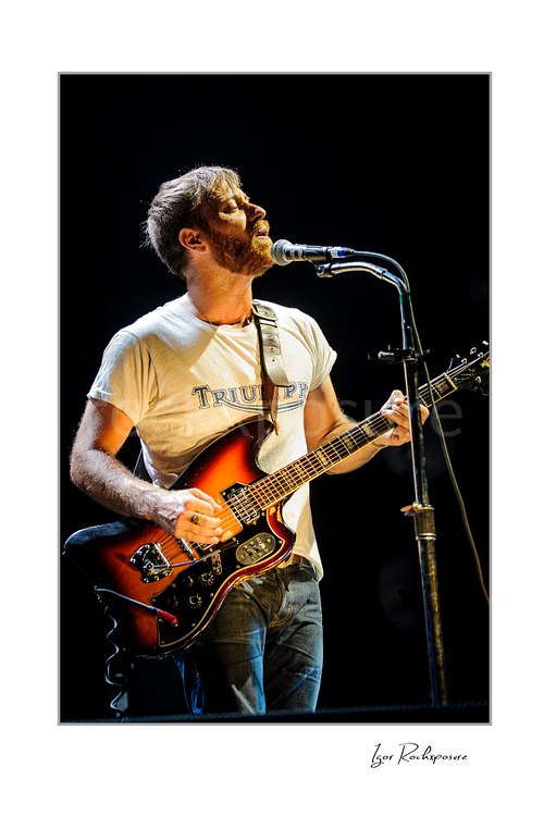 Vertical color image of Dan Auerbach of The Black Keys singing into a microphone while playing guitar under warm stage lighting