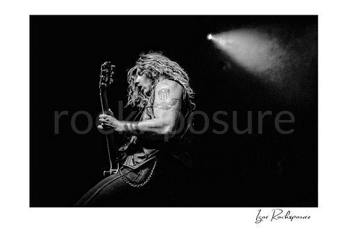 Horizontal black and white image of Jared James Nichols playing guitar onstage with flying hair under a bright spotlight