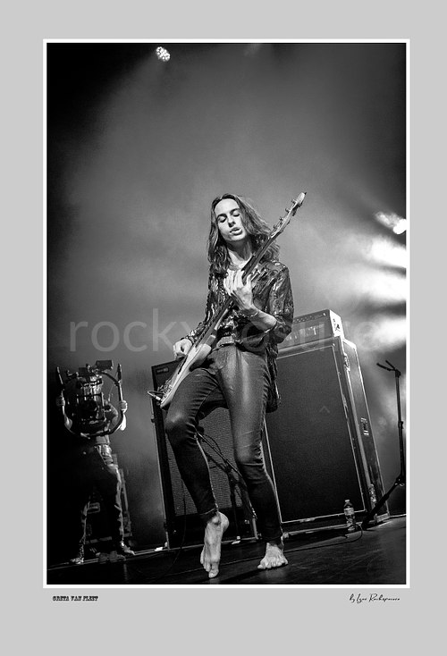 Vertical black and white image of Greta Van Fleet bassist Sam Kiszka performing live with his bass guitar on stage