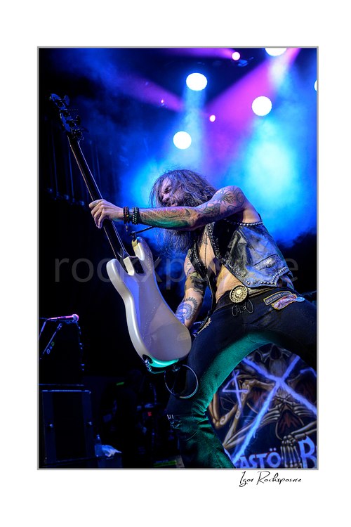 ertical color image of Jan LeGrow of Beasto Blanco playing guitar in a dramatic close-up, with long hair in motion and vivid stage lights glowing behind him