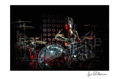 Horizontal color image of Tommy Lee performing at the drum kit during a Mötley Crüe concert, framed by cymbals with dramatic red stage light