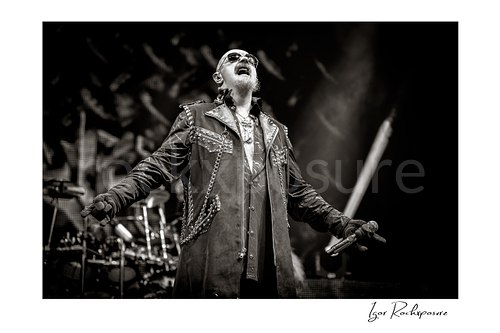 Horizontal black and white image of Rob Halford of Judas Priest performing live on stage, holding a microphone with his arms extended and the drum kit visible in the background
