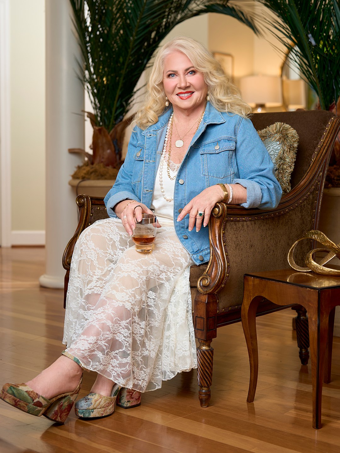 A blonde woman sits in an elegant hair wearing a lace skirt and blue denim jacket while smiling and looking towards the camera