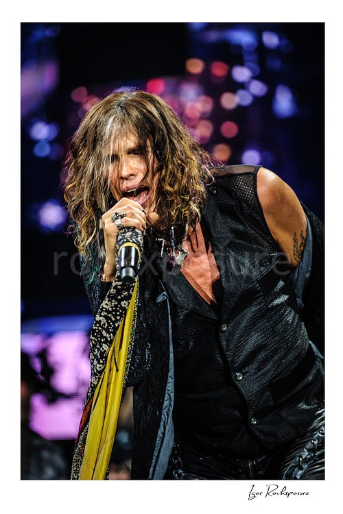 Vertical color image of Steven Tyler of Aerosmith performing live on stage with a handheld microphone, scarf-wrapped mic stand, and tousled hair against colorful blurred lights