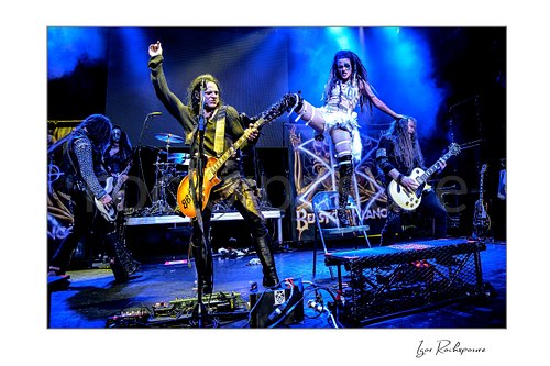 Horizontal color image of Beasto Blanco performing live on stage, with Chuck Garric at the microphone, Calico Cooper posed on a platform, and Jan LeGrow and Chris Latham playing guitar under vivid blue stage light