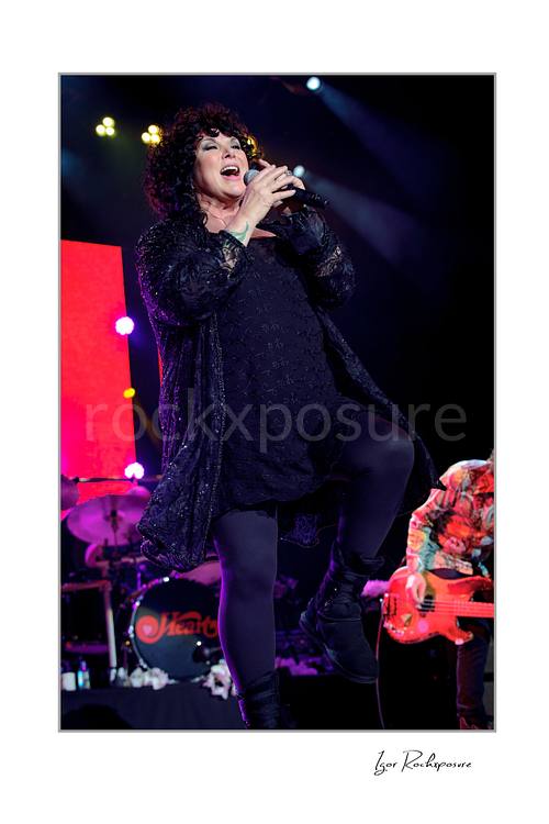 Vertical color image of Ann Wilson singing into a microphone during a Heart concert, lifting one leg in a dynamic stage pose under vivid purple and blue light