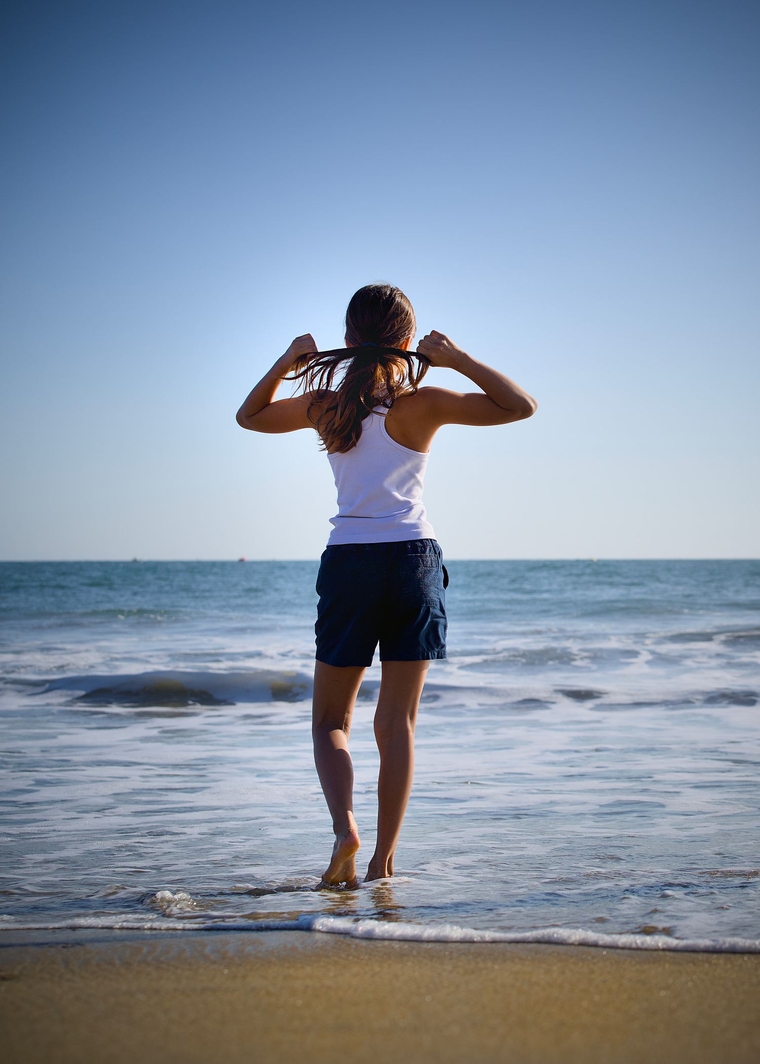 Child standing at the edge of the ocean while tying her hair before a portrait session.