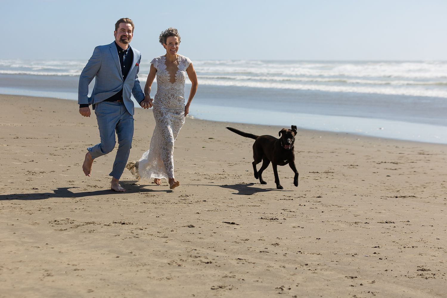 A newlywed couple running along the beach with their dog