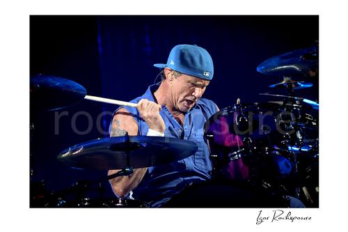 Horizontal color image of Chad Smith of Red Hot Chili Peppers playing drums in a focused live performance close-up, with drumstick raised above a purple drum kit under deep blue stage light