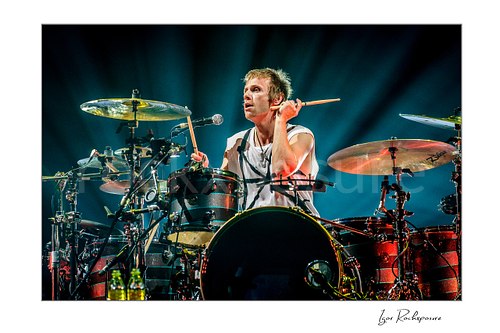 Horizontal color image of Dominic Howard of Muse playing drums with one stick held beside his head under radiant blue stage beams