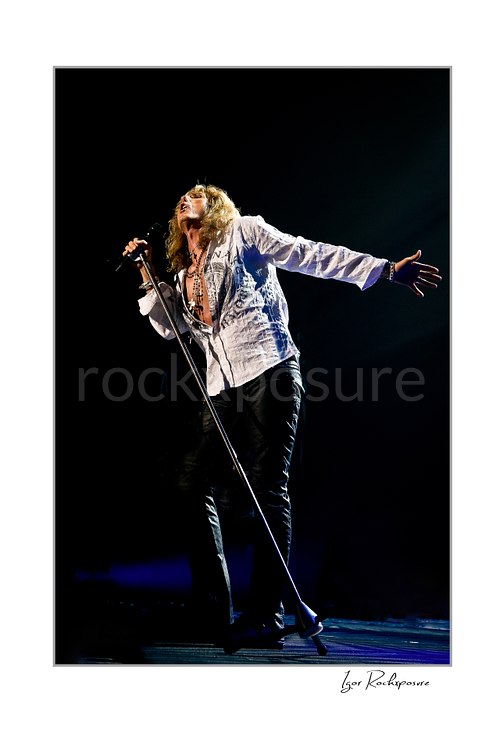 Vertical color image of David Coverdale of Whitesnake performing live on stage, singing into a microphone while extending one arm under dramatic concert lighting