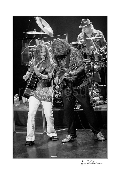 Vertical black and white image of Jon Davison, Billy Sherwood, and Alan White of Yes performing onstage together with acoustic guitar, bass, and drum kit