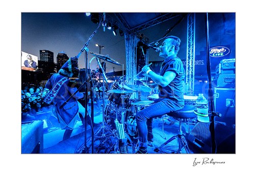 Horizontal color image of Stig Pedersen and Laust Sonne performing on stage in blue concert light, with Laust Sonne singing at the drum kit and a crowd visible beyond the stage