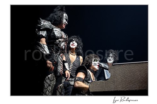 Horizontal color photograph of the full KISS band in full makeup and costumes gathered closely around Eric Singer as he plays piano and sings in a theatrical staged moment