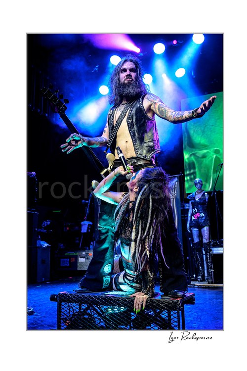 Vertical color image of Calico Cooper and Jan LeGrow of Beasto Blanco performing live on stage, with Cooper kneeling and singing below while LeGrow stands above her with arms spread wide under vivid blue and magenta stage lights