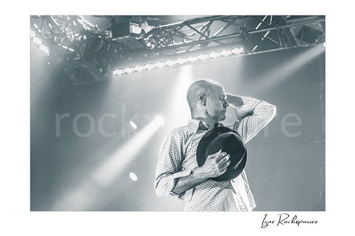 Horizontal black and white image of Gord Downie of The Tragically Hip in an emotional live performance moment, holding his hat to his chest with one hand behind his neck under soft stage light