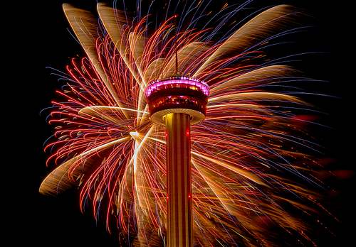 A horizontal fine art long exposure photograph of vibrant New Year's Eve fireworks exploding symmetrically around the historic Tower of the Americas in San Antonio, Texas.