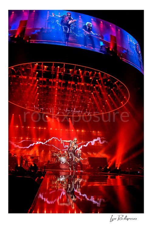 Vertical color image of Queen performing onstage beneath a massive circular lighting rig and giant overhead screen under vivid red concert light