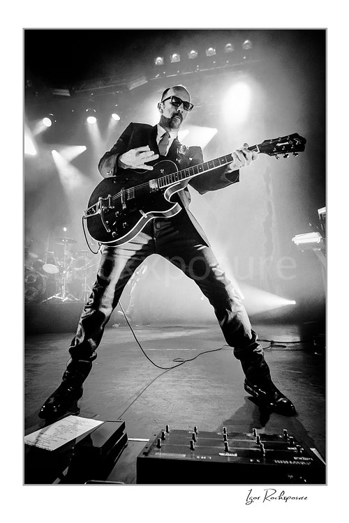 Vertical black and white image of Duke Erikson performing with electric guitar during a Garbage concert in a dramatic low-angle stage view under bright backlight