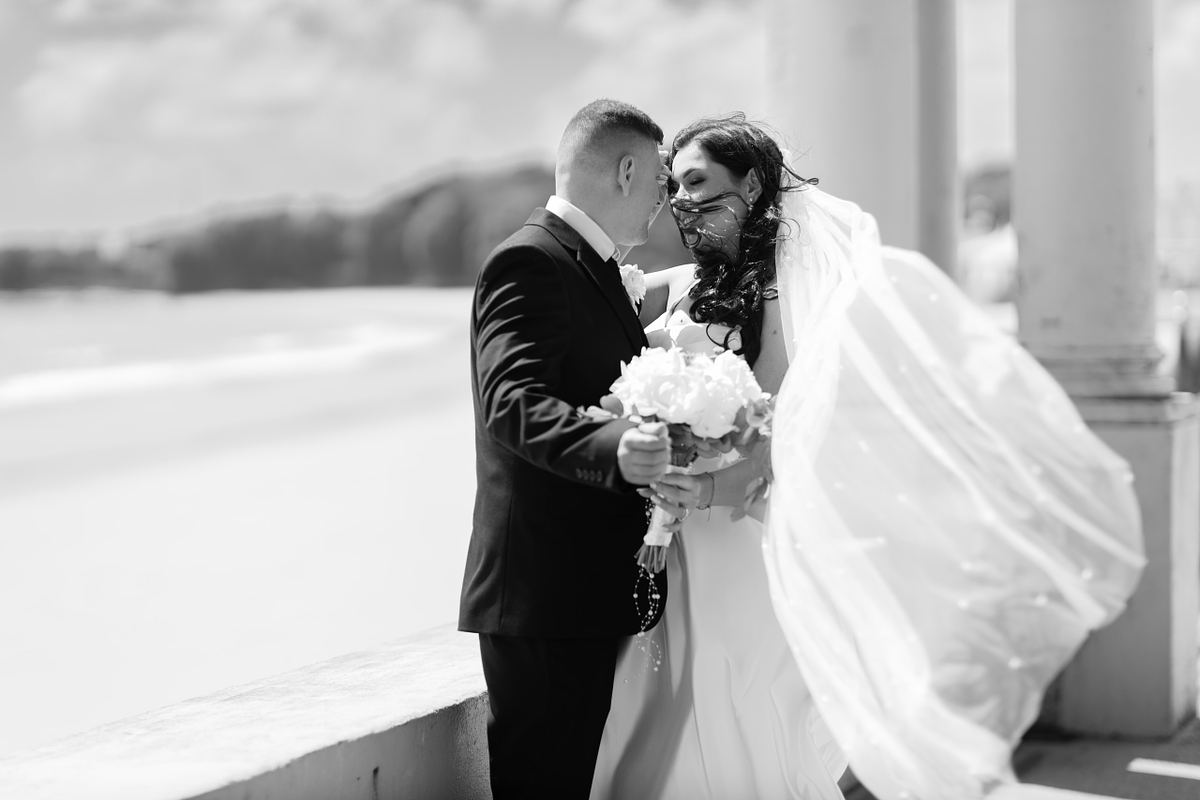 Black and white portrait of bride and groom with wind-blown veil by the sea at Barry Island, photographed by Marta Vershynina South Wales wedding photographer