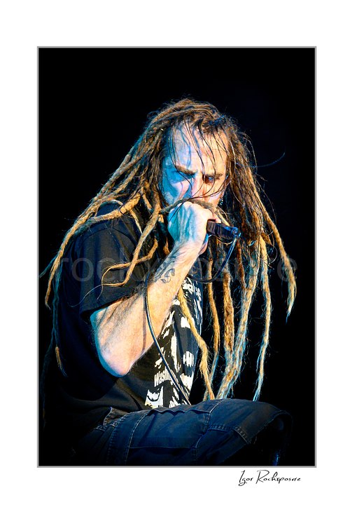 Vertical color image of Randy Blythe of Lamb of God in a tight stage close-up singing into a handheld microphone with long dreadlocks