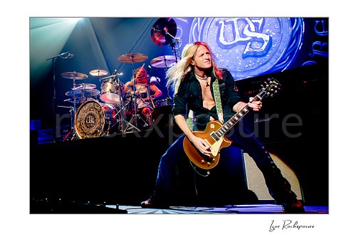 Horizontal color image of Doug Aldrich of Whitesnake performing live on stage, with Aldridge at left behind the drum kit and Aldrich at right playing guitar under bright blue concert lighting