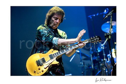 Horizontal color image of Steve Hackett performing live with electric guitar against a blue stage background