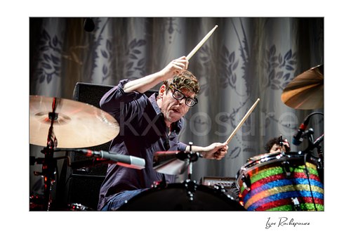 Horizontal color image of Patrick Carney of The Black Keys raising drumsticks at the drum kit in front of a patterned stage curtain