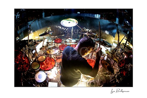 Horizontal color image of Korn drummer Ray Luzier seen from behind at the drums with one drumstick raised above the stage and crowd