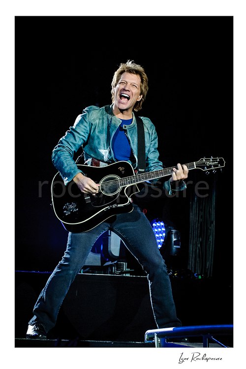 Vertical color image of Jon Bon Jovi performing live with a black acoustic guitar, smiling broadly on stage with blue concert lights behind him