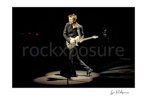 Horizontal color image of Matt Bellamy of Muse playing guitar alone in a spotlight on a dark stage