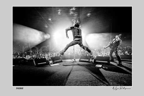 Horizontal black and white image of Bret Michaels leaping in midair and C.C. DeVille performing with electric guitar during a Poison concert in a dramatic backlit stage scene