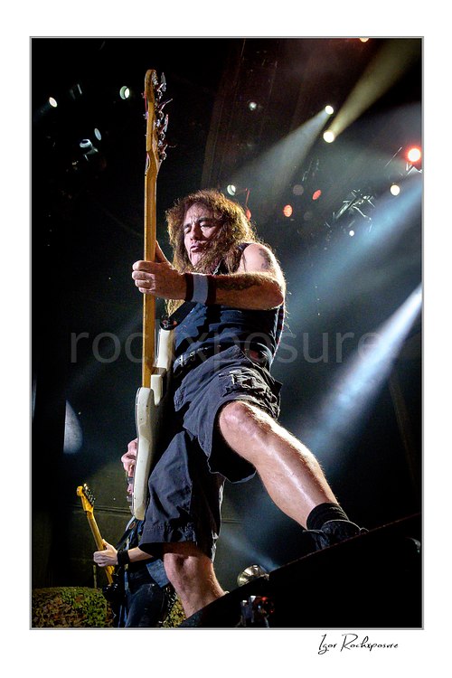 Steve Harris of Iron Maiden performing live on stage in a dramatic low-angle shot while playing bass guitar under bright concert lights.