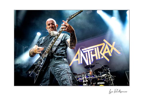 Horizontal color image of Scott Ian of Anthrax performing live on stage, playing guitar in front of the Anthrax stage backdrop under bright concert lighting