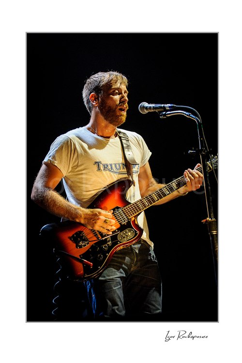 Vertical color image of Dan Auerbach of The Black Keys singing into a microphone while playing guitar under warm stage lighting