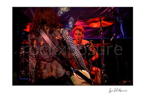 Horizontal color image of Jan LeGrow and Sean Sellers of Beasto Blanco performing live on stage, with LeGrow seen from behind in the foreground and Sellers at the drum kit under vivid red stage light